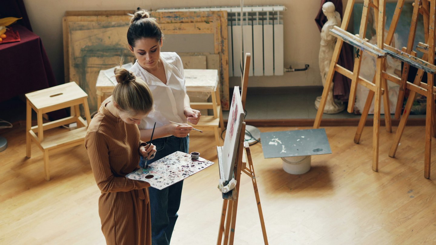 Un istruttore d'arte guida uno studente che dipinge in uno studio luminoso.